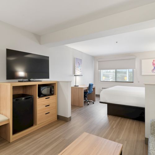 A modern hotel room with a king-size bed, TV, desk, chair, mini-fridge, and microwave. The room has wooden flooring and minimalist decor.