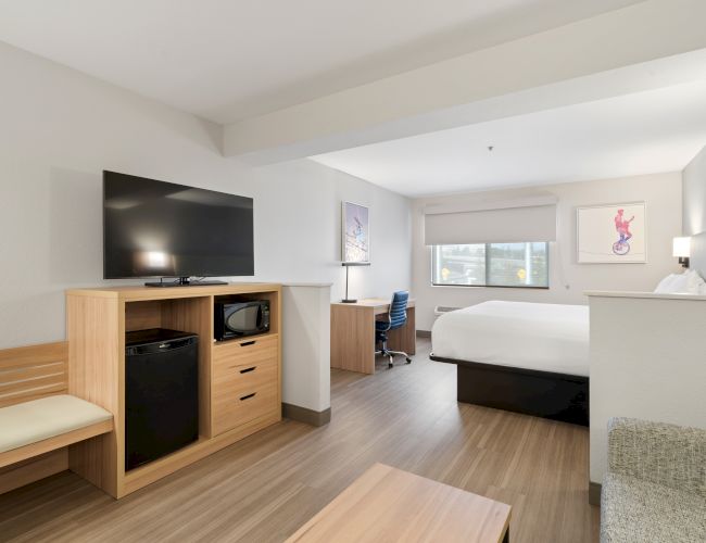 A modern hotel room with a king-size bed, TV, desk, chair, mini-fridge, and microwave. The room has wooden flooring and minimalist decor.