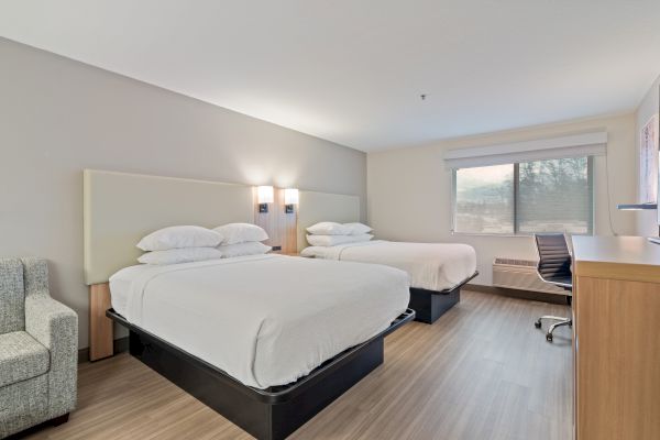 A modern hotel room featuring two double beds, an armchair, a desk with a chair, a large window with a view, and minimalistic decor.