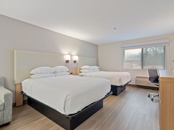 A modern hotel room featuring two double beds, an armchair, a desk with a chair, a large window with a view, and minimalistic decor.