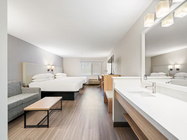 A modern hotel room with two beds, a sofa, a coffee table, a long countertop with a mirror, and ample lighting. The decor is minimalist.