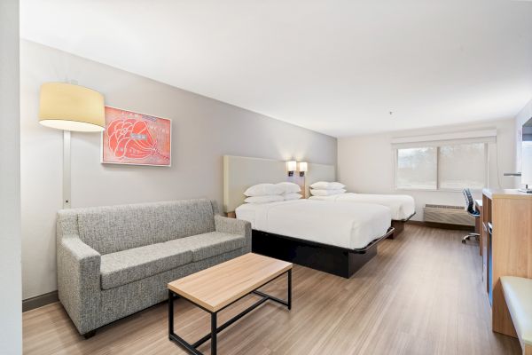 This image shows a hotel room with two double beds, a gray sofa, a small coffee table, a bright floor lamp, and a desk. The walls are light-colored.