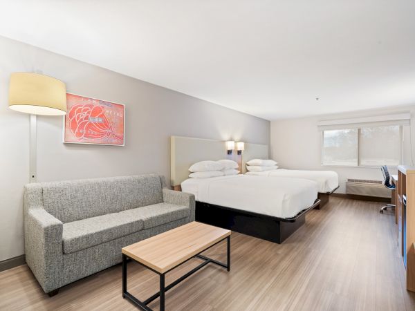 This image shows a hotel room with two double beds, a gray sofa, a small coffee table, a bright floor lamp, and a desk. The walls are light-colored.