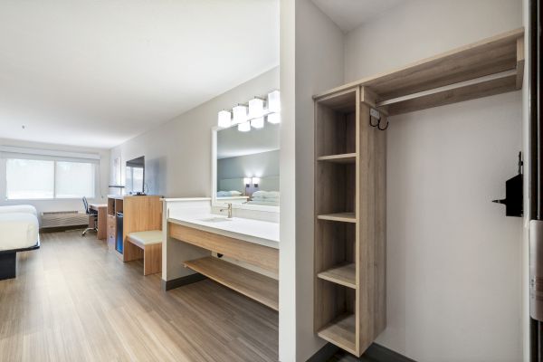 A modern, well-lit hotel room featuring a bed, desk, TV, vanity area with mirror, and an open closet with shelves and hangers.