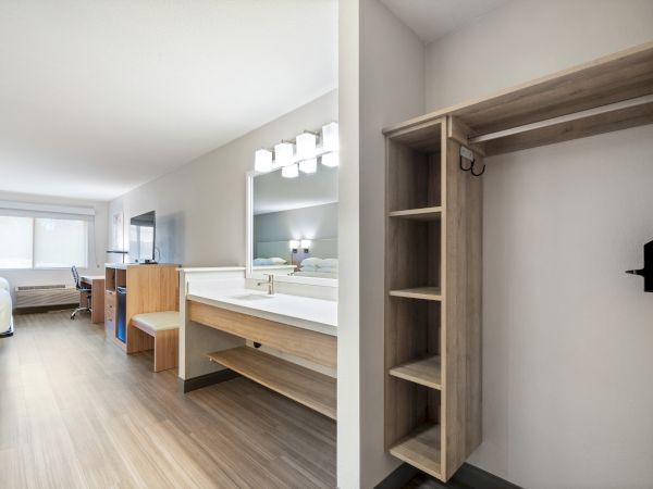 A modern, well-lit hotel room featuring a bed, desk, TV, vanity area with mirror, and an open closet with shelves and hangers.