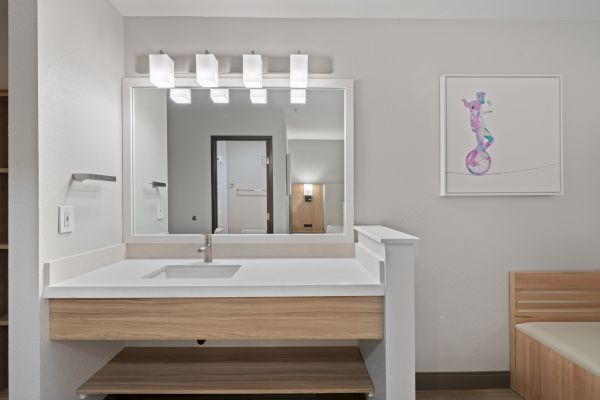 A modern bathroom with a large mirror, sink with a wooden vanity, bright lighting, and a colorful framed artwork on the wall.