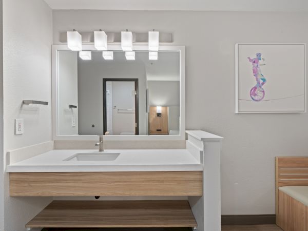 A modern bathroom with a large mirror, sink with a wooden vanity, bright lighting, and a colorful framed artwork on the wall.