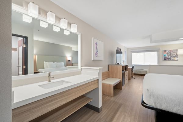 The image shows a modern hotel room with a bed, a vanity area, a TV, desk, chair, and window, decorated in a minimalist style with soft colors.