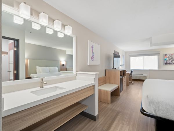 The image shows a modern hotel room with a bed, a vanity area, a TV, desk, chair, and window, decorated in a minimalist style with soft colors.