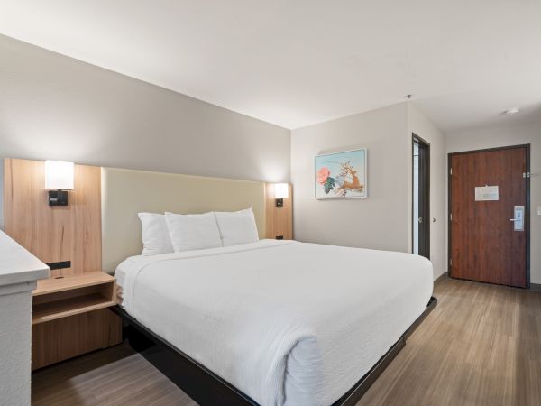 A modern hotel room features a large bed with white linens, wooden headboard, wall art, bedside lamps, and a door leading to a bathroom.