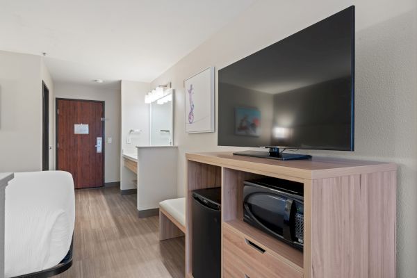 A hotel room with a TV, a microwave, a mini fridge, a bed, a sink, and a bathroom door. The room is simple and modern in design.