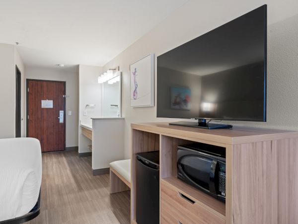 A hotel room with a TV, a microwave, a mini fridge, a bed, a sink, and a bathroom door. The room is simple and modern in design.
