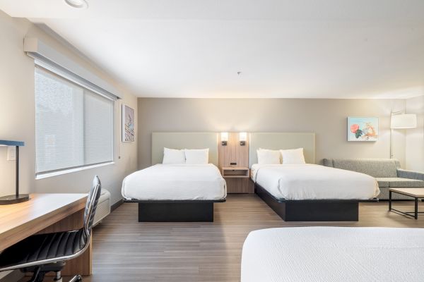 A clean, modern hotel room with two beds, a work desk, a chair, a sofa, and artwork on the walls.