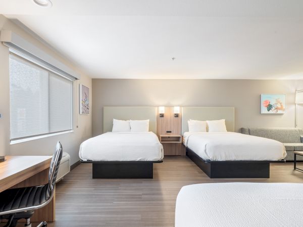 A clean, modern hotel room with two beds, a work desk, a chair, a sofa, and artwork on the walls.