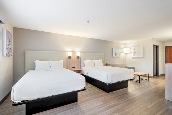 This image shows a modern hotel room with two queen-sized beds, minimalist decor, wall art, wooden flooring, and soft lighting.