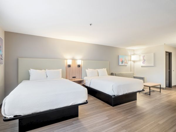 This image shows a modern hotel room with two queen-sized beds, minimalist decor, wall art, wooden flooring, and soft lighting.