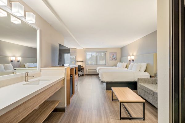 This image shows a modern hotel room with two beds, a sofa, a large mirror, a coffee table, and a TV unit. The room is simply furnished and clean.