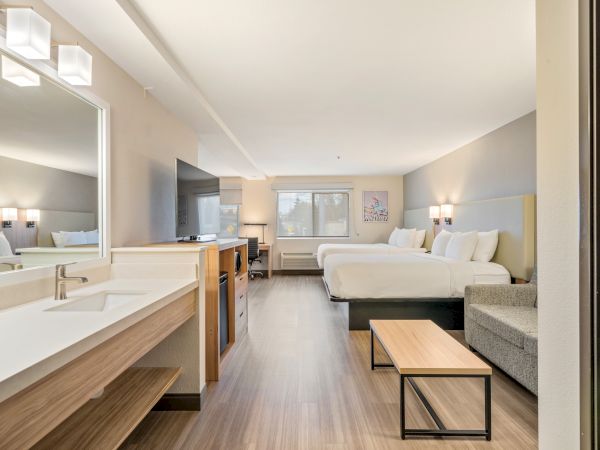 This image shows a modern hotel room with two beds, a sofa, a large mirror, a coffee table, and a TV unit. The room is simply furnished and clean.