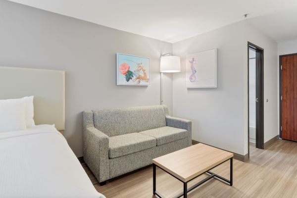 A modern hotel room features a bed, a gray sofa, a wooden coffee table, two wall artworks, a floor lamp, and a doorway leading to a bathroom.