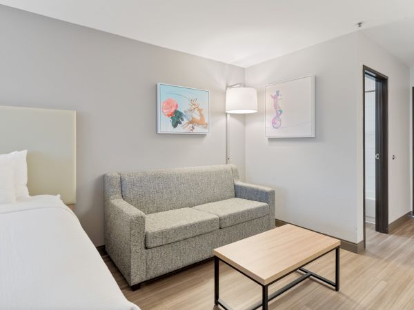 A modern hotel room features a bed, a gray sofa, a wooden coffee table, two wall artworks, a floor lamp, and a doorway leading to a bathroom.