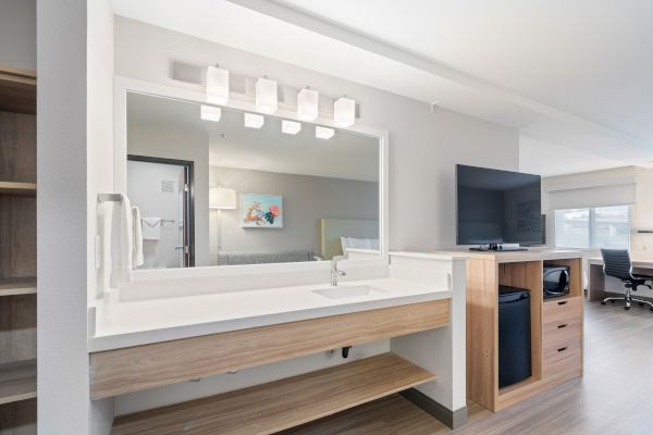 The image shows a modern hotel room with a vanity sink, well-lit mirror, a TV, a mini-fridge, and a desk with a chair in the background.