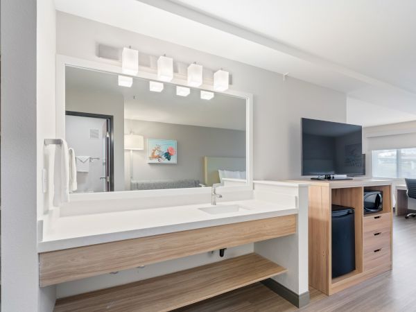 The image shows a modern hotel room with a vanity sink, well-lit mirror, a TV, a mini-fridge, and a desk with a chair in the background.