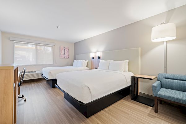 A modern hotel room with two beds, a desk, a chair, and a floor lamp. The room is well-lit, featuring a large window.