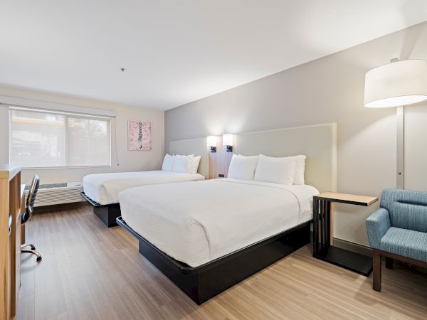 A modern hotel room with two beds, a desk, a chair, and a floor lamp. The room is well-lit, featuring a large window.