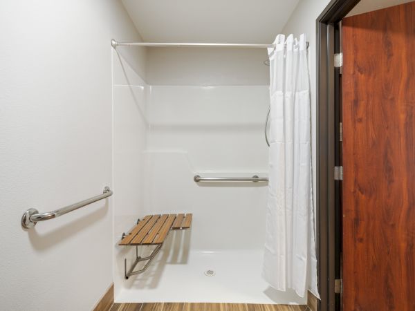 This is an accessible shower with handrails, a wooden bench, and a white shower curtain, designed for ease of use.
