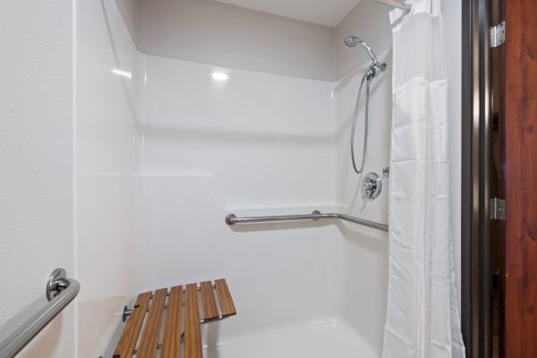 The image shows a walk-in shower with a wooden bench, grab bars, and a handheld showerhead, designed for accessibility.