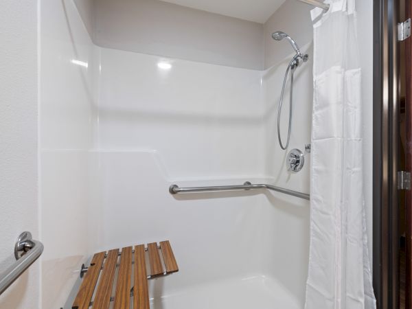 The image shows a walk-in shower with a wooden bench, grab bars, and a handheld showerhead, designed for accessibility.