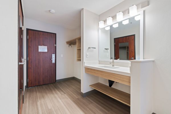 A modern bathroom area with a large mirror, sink, and shelf, next to an entryway with a wooden door and hanging space for clothes or luggage.