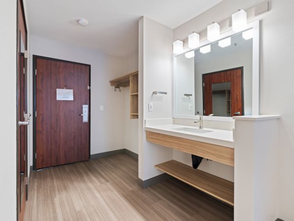 A modern bathroom area with a large mirror, sink, and shelf, next to an entryway with a wooden door and hanging space for clothes or luggage.