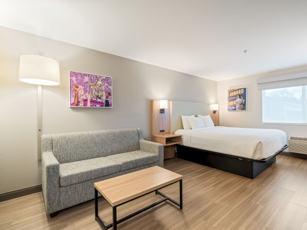 A modern hotel room features a bed, nightstands, a sofa, a coffee table, a floor lamp, wall art, and a large window with natural light.