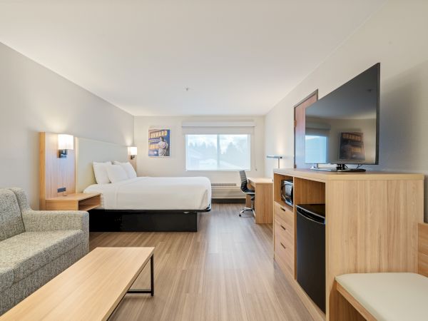 A modern hotel room with a bed, flatscreen TV, sofa, desk, and ample natural light from a window. The room has a clean and minimalist design.