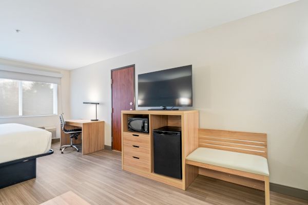 The image shows a hotel room featuring a bed, desk with chair, lamp, TV on a stand with a microwave, fridge, and a bench.