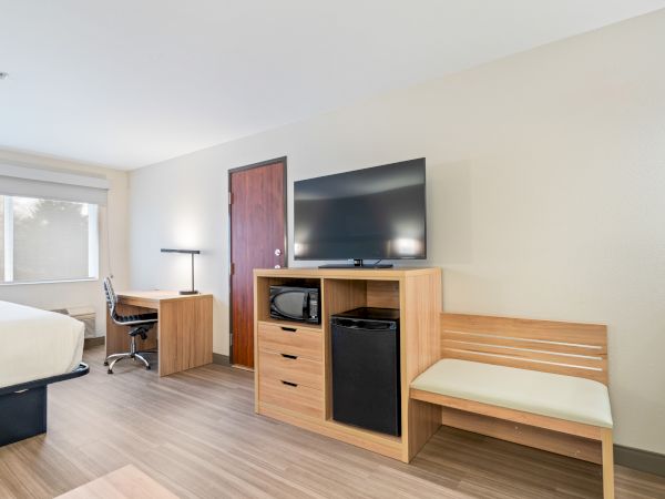The image shows a hotel room featuring a bed, desk with chair, lamp, TV on a stand with a microwave, fridge, and a bench.