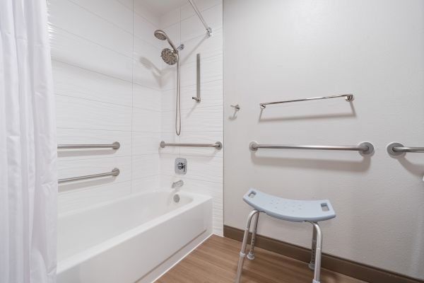 The image shows a bathroom with a white bathtub, multiple handrails, a showerhead, a shower curtain, and a shower chair.