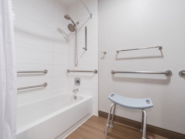 The image shows a bathroom with a white bathtub, multiple handrails, a showerhead, a shower curtain, and a shower chair.