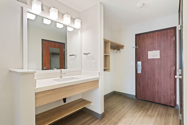 The image shows a modern entryway or bathroom area with a wooden door, a sink with a large mirror, and shelves. The floor is hardwood.