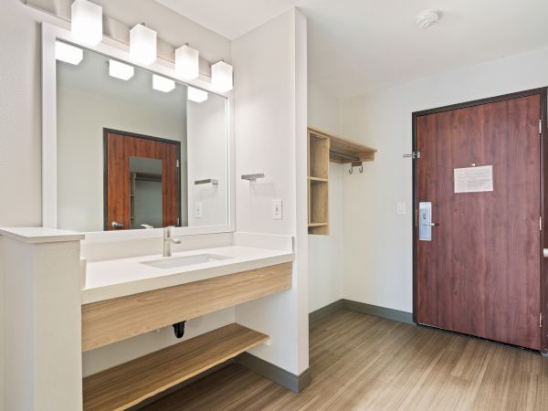 The image shows a modern entryway or bathroom area with a wooden door, a sink with a large mirror, and shelves. The floor is hardwood.