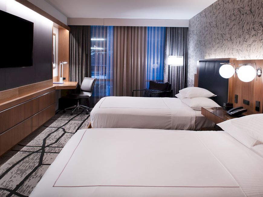 A modern hotel room with two neatly made beds, a flat-screen TV, a work desk with a chair, and large windows with curtains ending the sentence.