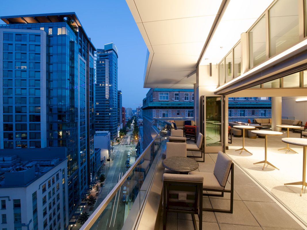 A modern balcony with seating overlooks a cityscape featuring high-rise buildings and a street stretching into the distance near dusk.