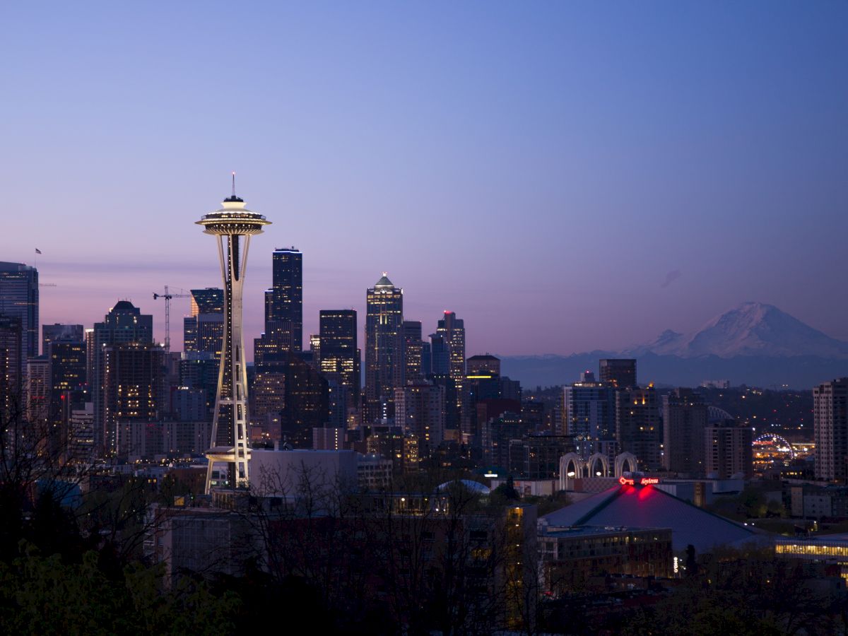 This is a cityscape of Seattle at dusk, featuring the Space Needle prominently and Mount Rainier in the background, with buildings illuminated.
