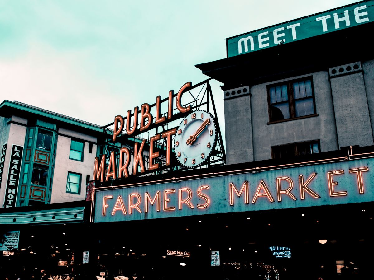 The image shows the entrance to a public farmers market with neon signs and a large clock, set against the backdrop of nearby buildings and an overcast sky.