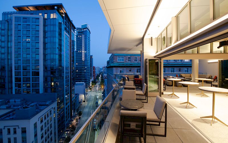 A modern cityscape at twilight, featuring tall buildings and a spacious balcony with seating and tables overlooking the urban scene.